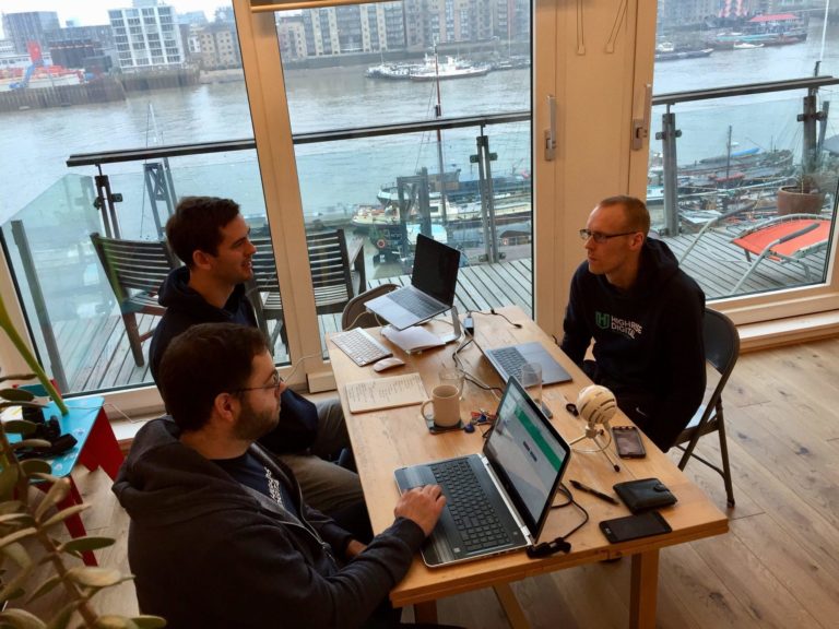 3 adults males sat around a small table discussing work, in front of a backdrop of the river Thames.